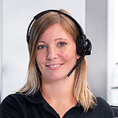 Eine Frau mit blonden Haaren, die ein schwarzes Headset und ein schwarzes Hemd trägt, lächelt in die Kamera und scheint sich in einer Kundendienst- oder Call-Center-Umgebung zu befinden.