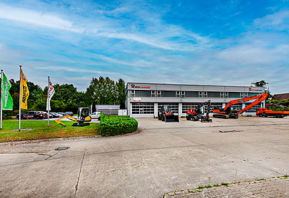 Ein Geschäftsgebäude mit großen Garagentoren und mehreren Baufahrzeugen, darunter Bagger und Lader, die draußen geparkt sind. Auf dem Rasen sind Fahnen aufgestellt, und an der Seite sind Autos geparkt. Im Hintergrund sind Bäume zu sehen.