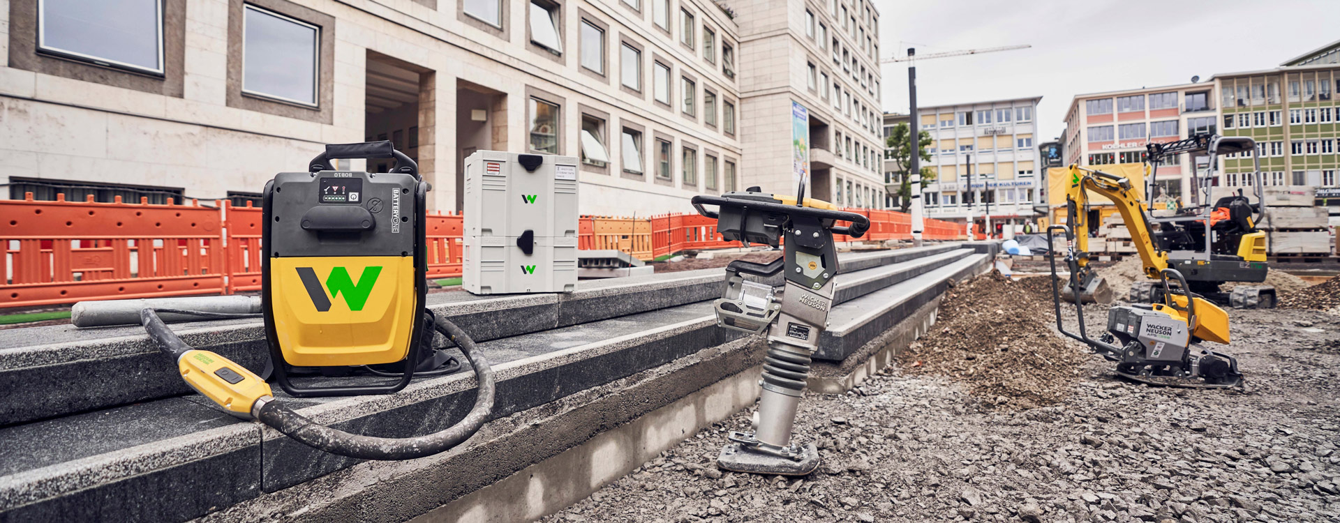 Baustelle mit verschiedenen gelben und grauen Geräten, Betonbarrieren und Gebäuden im Hintergrund