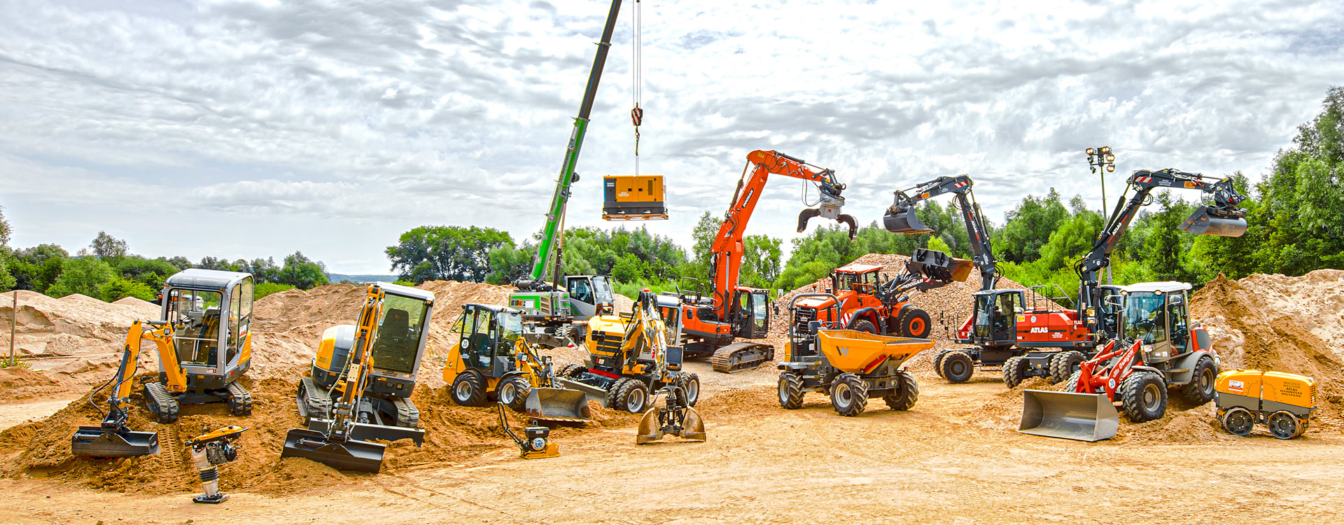 Eine Reihe von verschiedenen Baufahrzeugen und -geräten, darunter Bagger, Planierraupen, Lader, Kipper und ein Kran, sind auf einer unbefestigten Baustelle mit Sandhaufen und Bäumen im Hintergrund angeordnet.