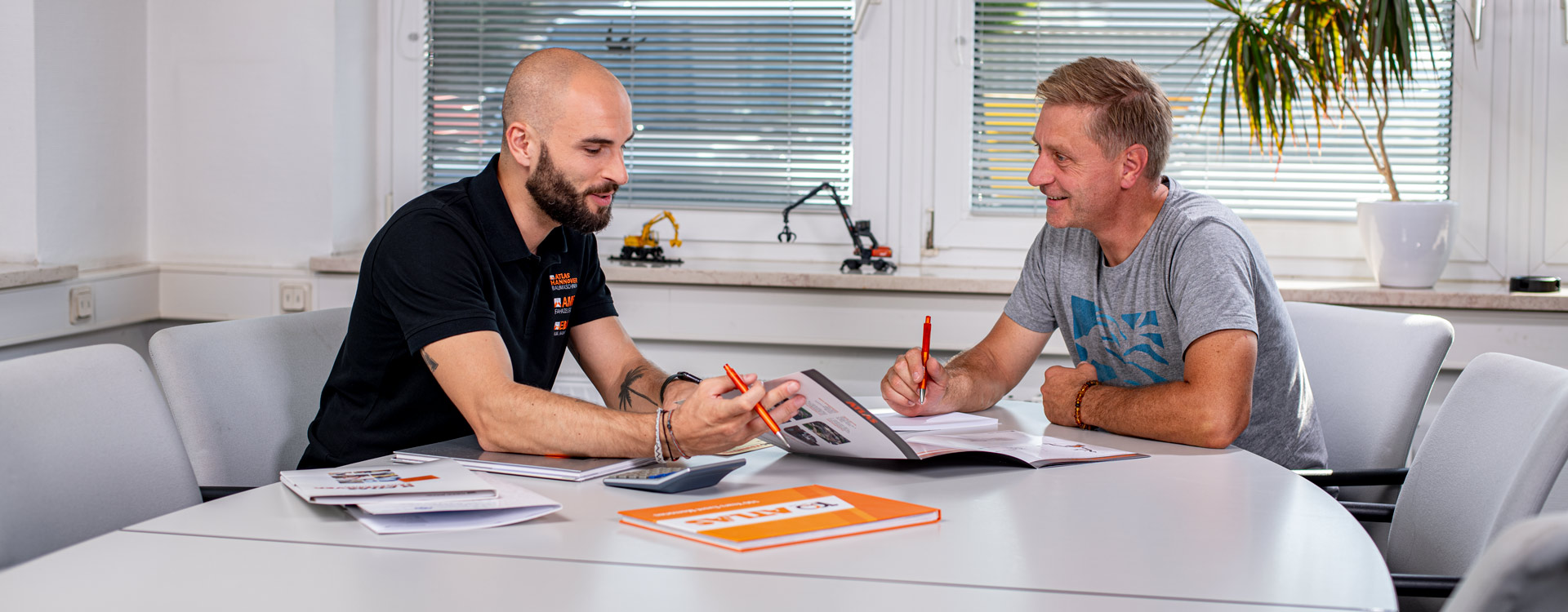 Zwei Männer sitzen an einem Tisch und besprechen Dokumente, jeder hält einen orangefarbenen Stift in der Hand. Einer trägt ein schwarzes Poloshirt, der andere ein graues T-Shirt. Sie lächeln, auf dem Tisch liegen Papiere, ein Taschenrechner und kleine Modellfahrzeuge.